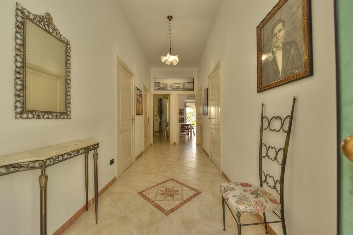 A hallway with a mirror, a chair, a framed picture, and a patterned floor.