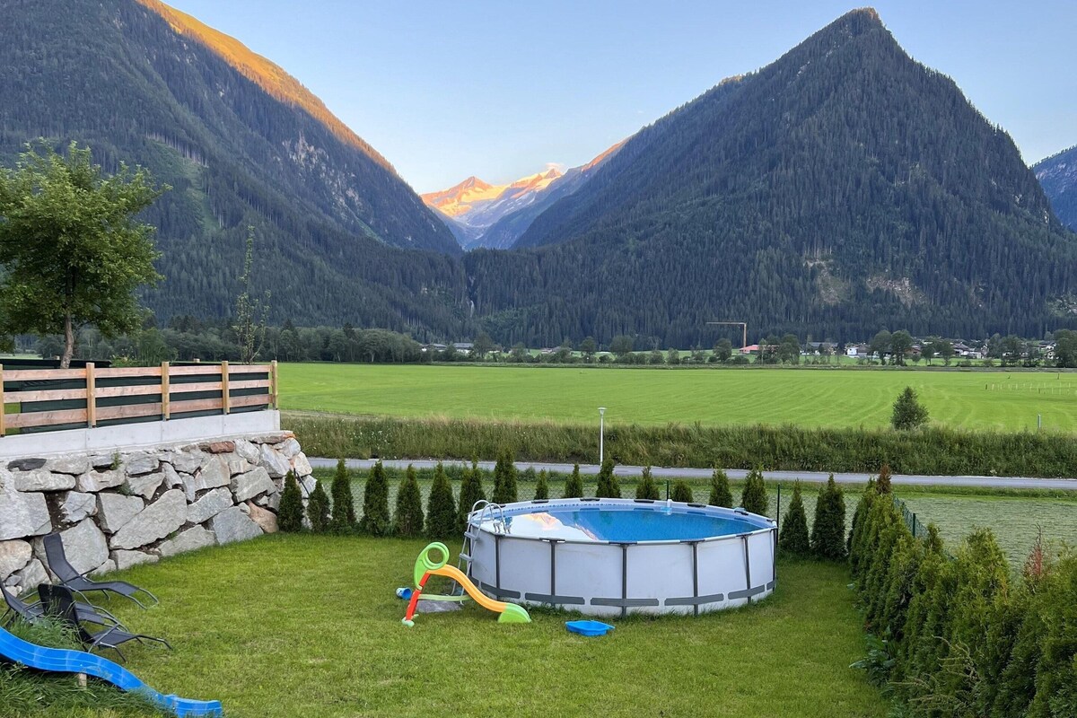 A wooden bench in a grassy area with a swimming pool and a playground, surrounded by mountains and a clear sky.