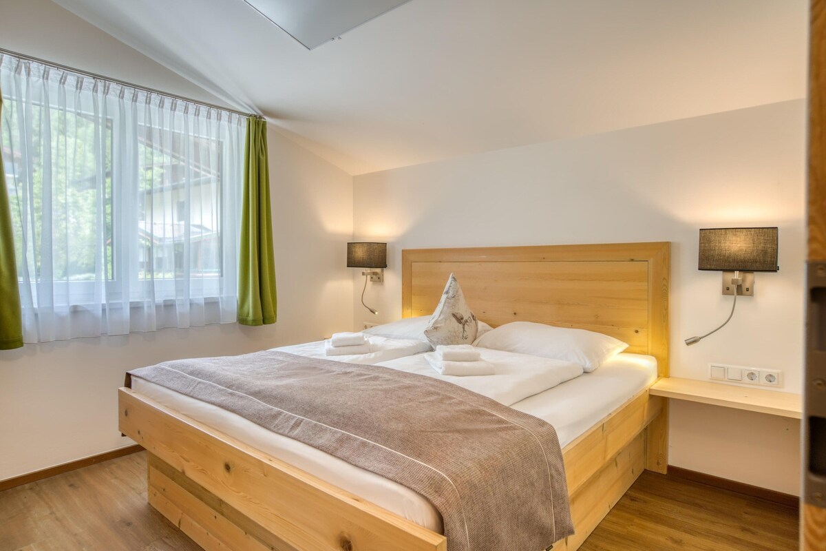 A wooden bed with a headboard, two bedside lamps, a bedside table, and a window with sheer curtains.