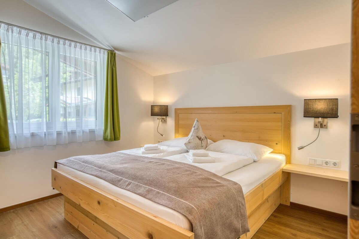 A neatly made bed with a wooden headboard, two bedside lamps, a bedside table, and a window with sheer curtains.