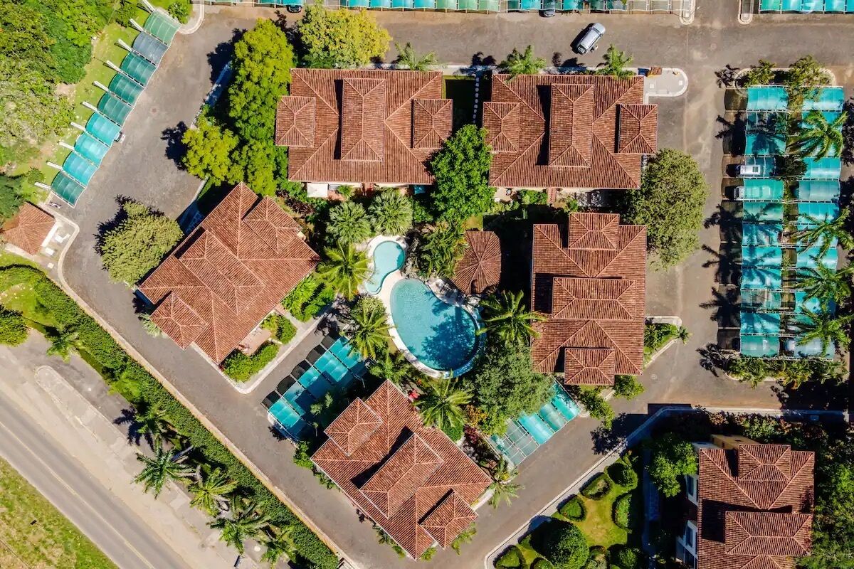 Vista aérea de un complejo turístico con varias edificaciones tipo bungalow, una piscina central y palmeras circundantes.