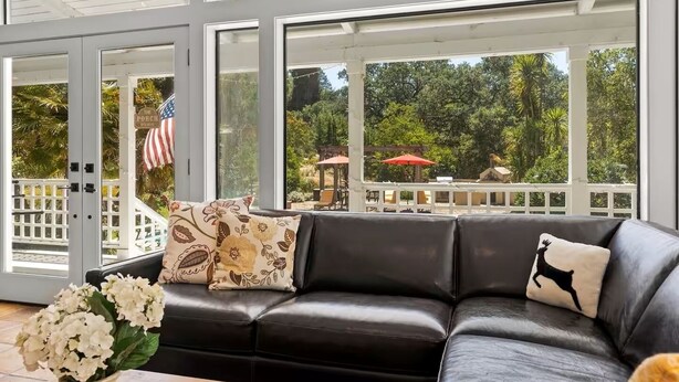Un canapé en cuir noir agrémenté de coussins à motifs floraux et animaliers, un drapeau américain blanc, et une vue sur une terrasse avec des meubles d’extérieur et de la verdure.