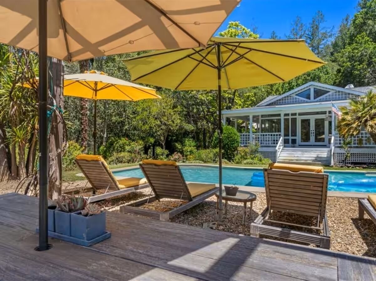 Um deck de madeira com espreguiçadeiras e guarda-sóis, uma piscina e uma casa ao fundo.