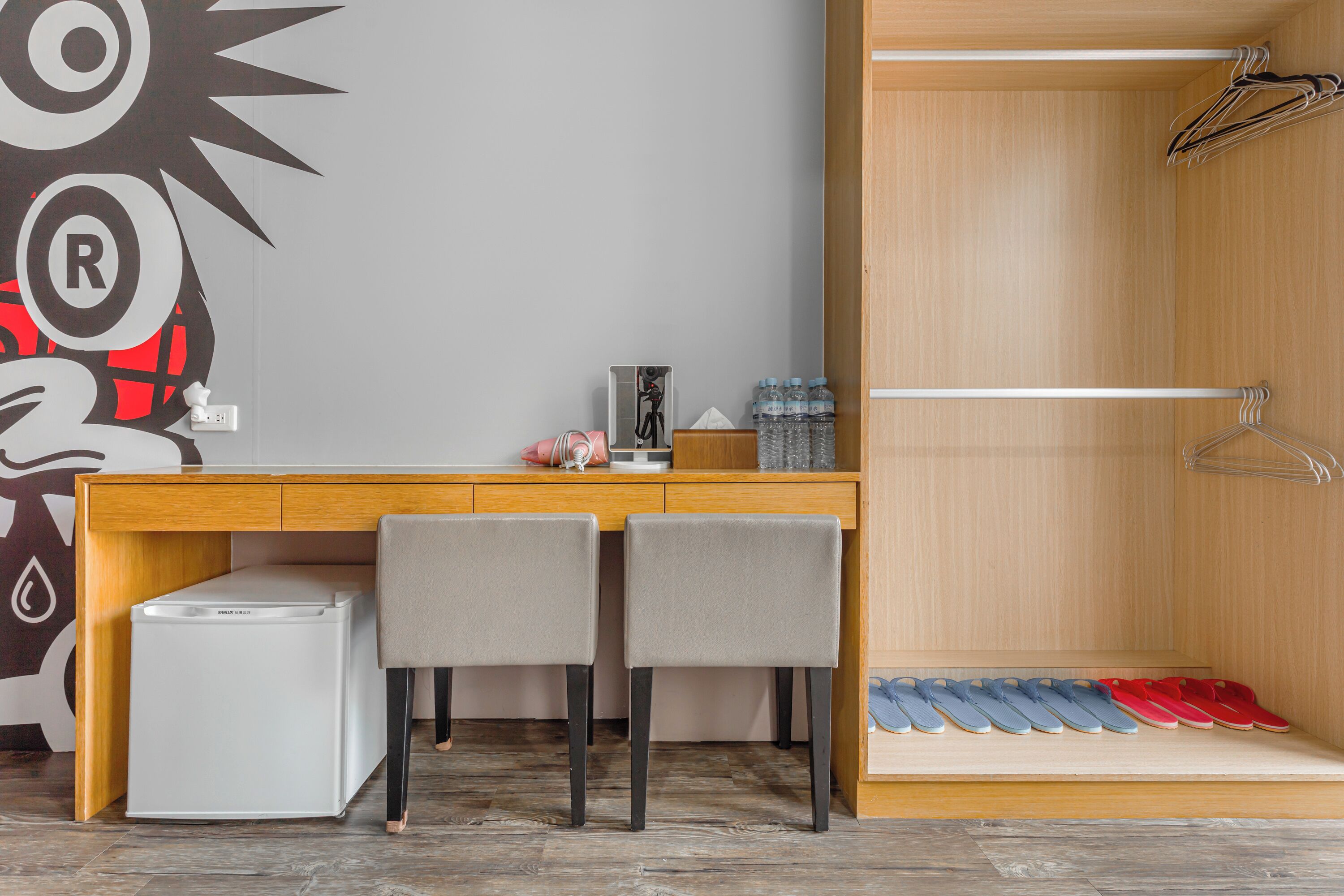 A compact room with a wooden desk, two chairs, a small refrigerator, a wall decoration, a water dispenser, and a shoe rack.