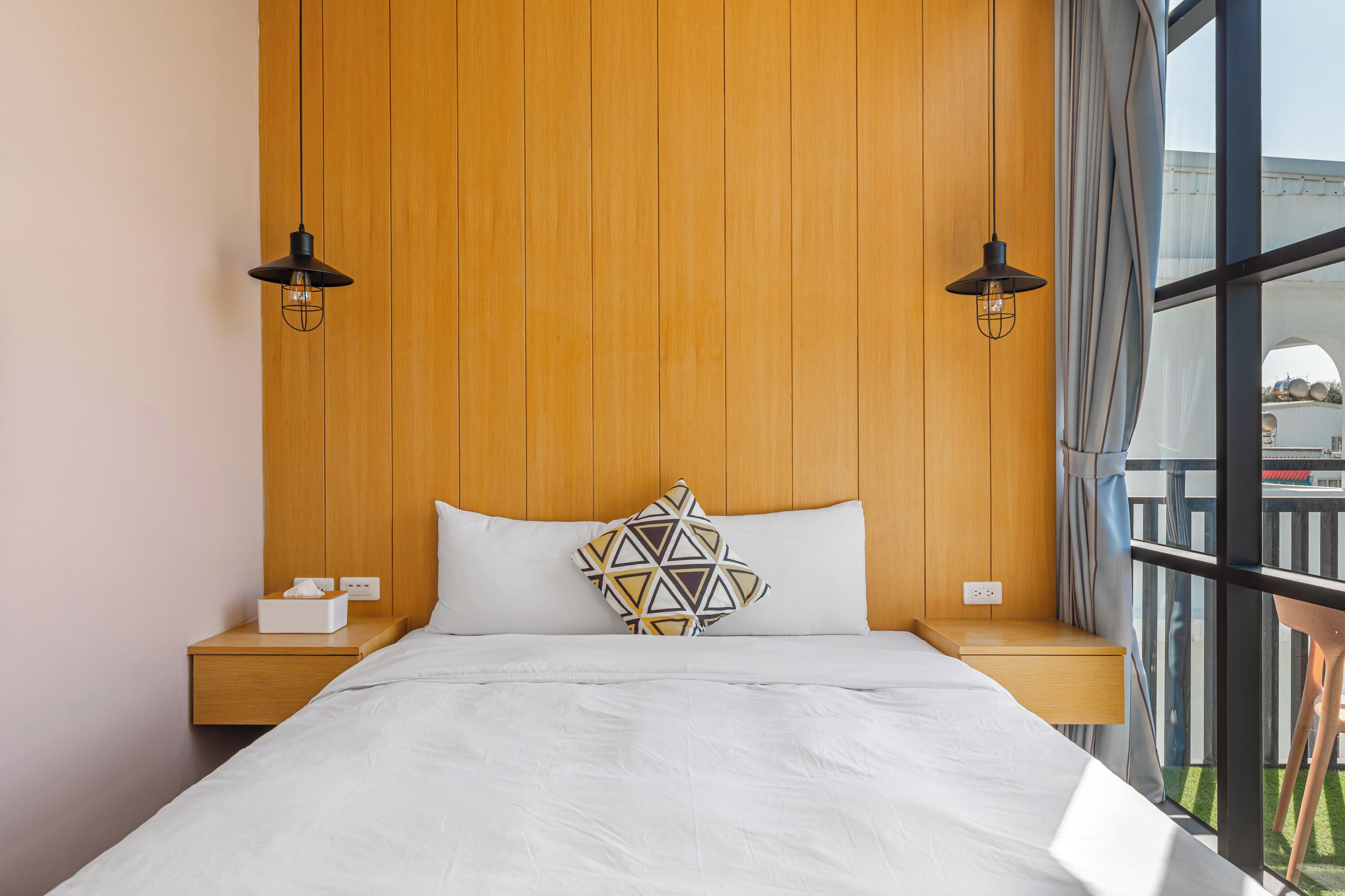 Habitación de hotel con cabecera de madera, cama con ropa blanca, almohada de estampado geométrico, dos mesitas de noche con lámparas y vistas amplias por ventanas grandes.