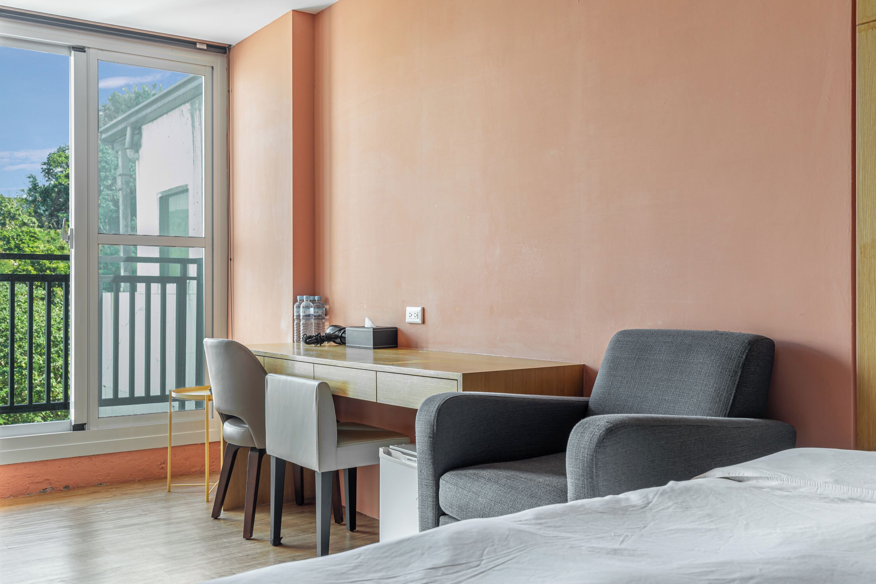 A room with a bed, a desk, and a chair. There is a window with a view of greenery outside.