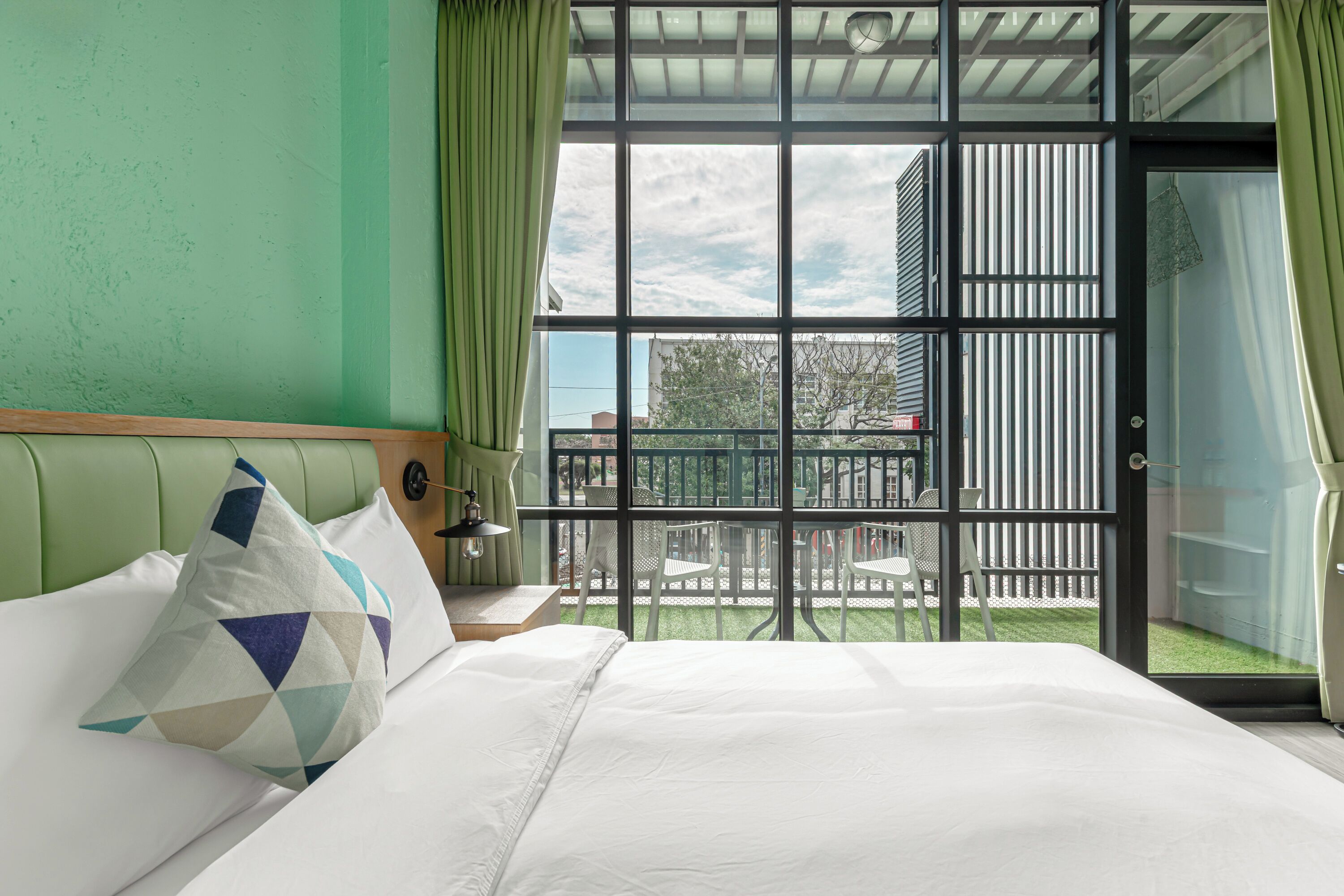 A hotel room with a bed, a nightstand, a lamp, and a view of the outside balcony.