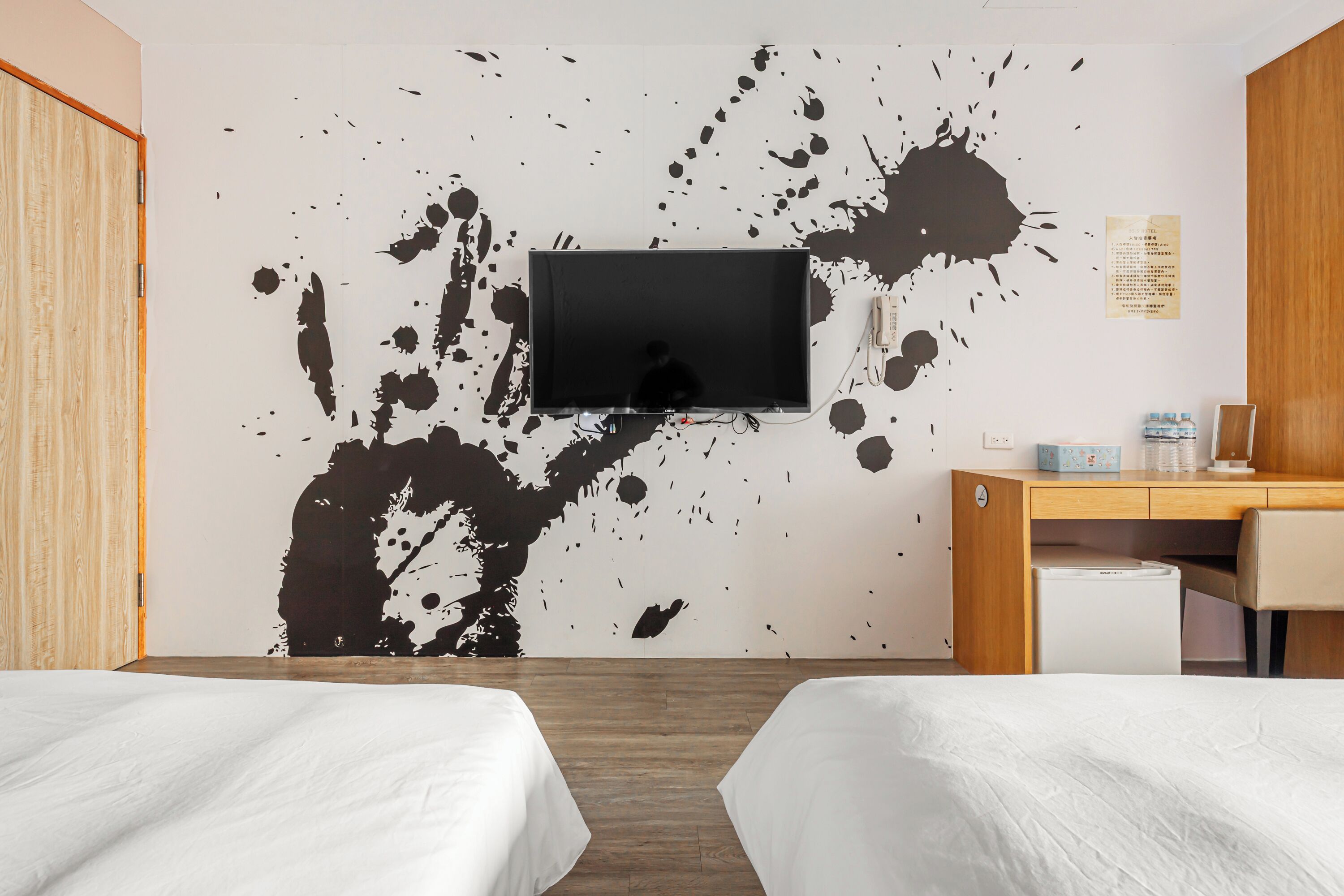 A hotel room with two beds, a TV mounted on the wall, a desk with a chair, and a small refrigerator.