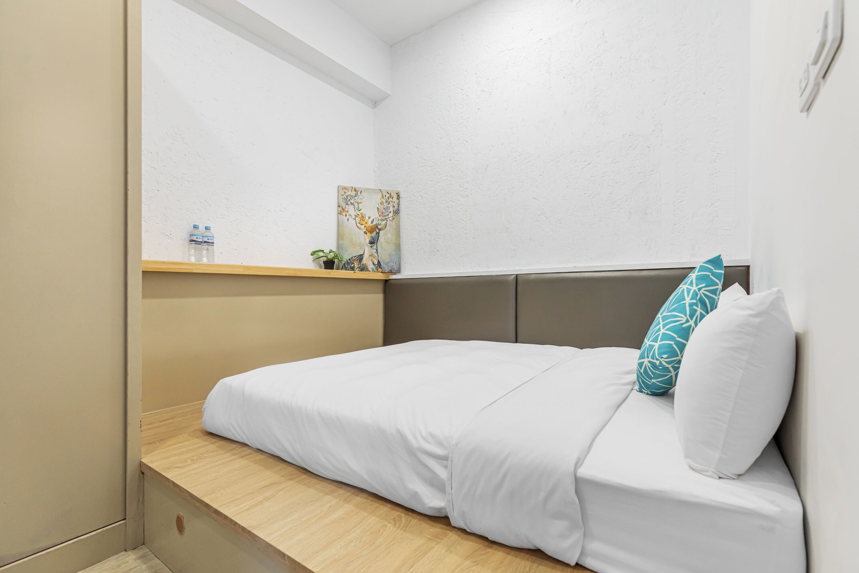A small, neatly made bed with white bedding and a teal pillow, a small shelf with a water bottle and a plant, and a painting of a deer on the wall.