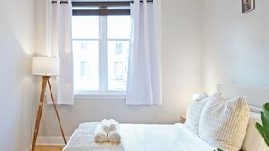 A hotel room with a bed, a bedside lamp, a window with curtains, and a view of buildings outside.