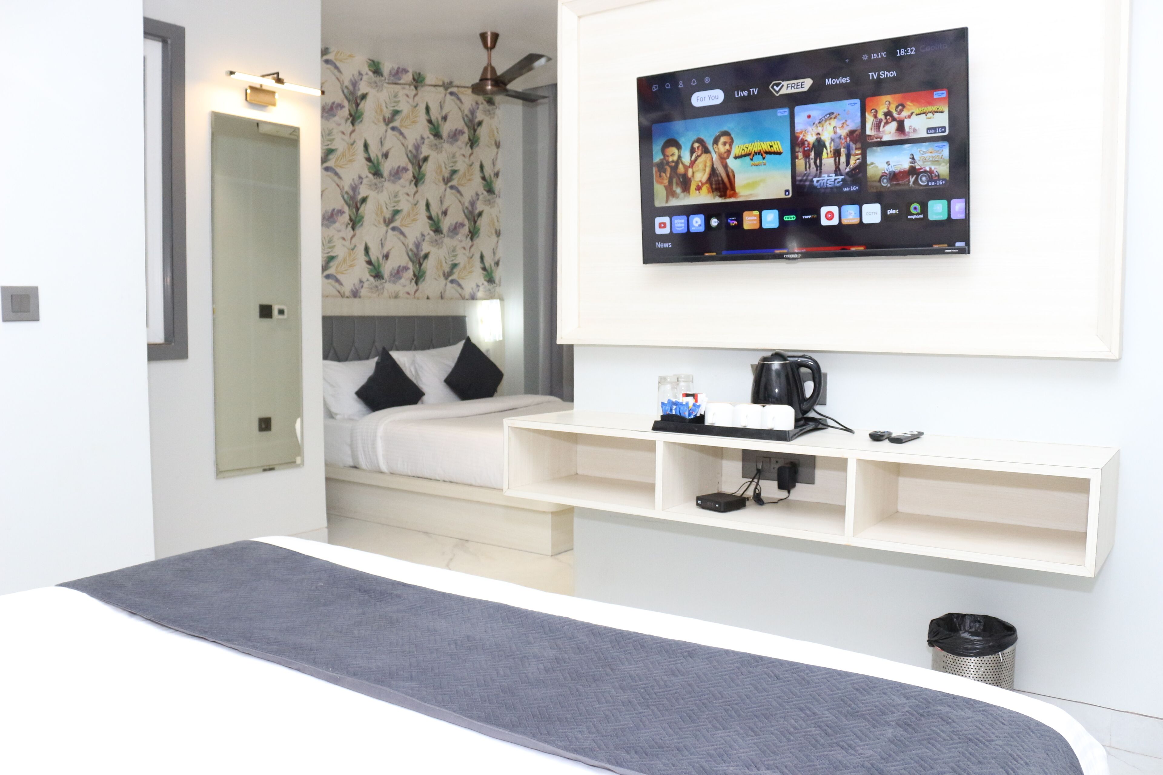 Moderni hotellihuone, jossa on suuri, seinälle asennettu litteänäyttöinen televisio, harmaalla päätyosalla varustettu sänky, valkoinen yöpöytä, jossa on vedenkeitin ja kaukosäätimet, sekä pieni roskakori.
