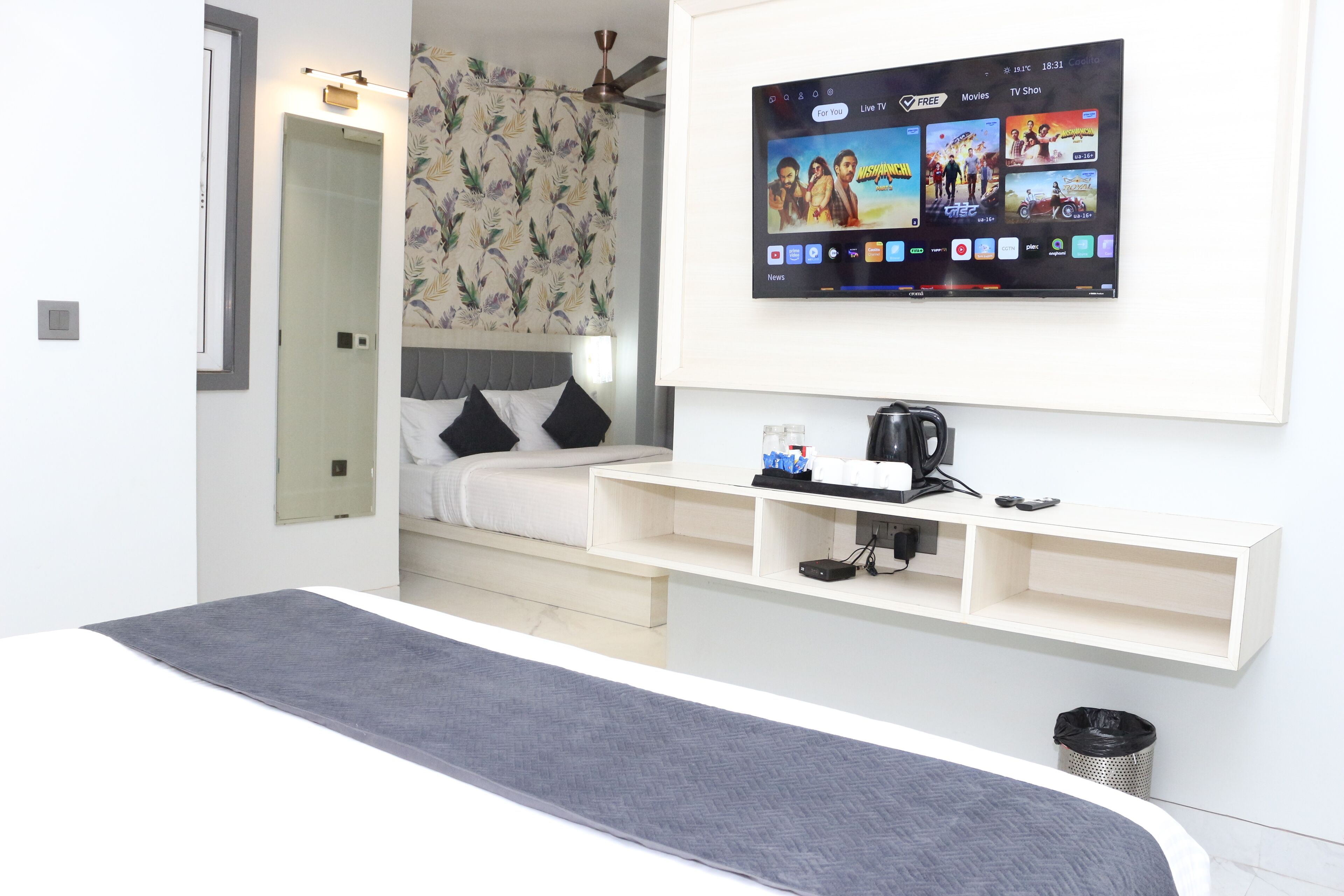 Une chambre d’hôtel moderne équipée d’un lit, d’un téléviseur fixé au mur, d’une petite étagère avec une bouilloire et d’une poubelle.