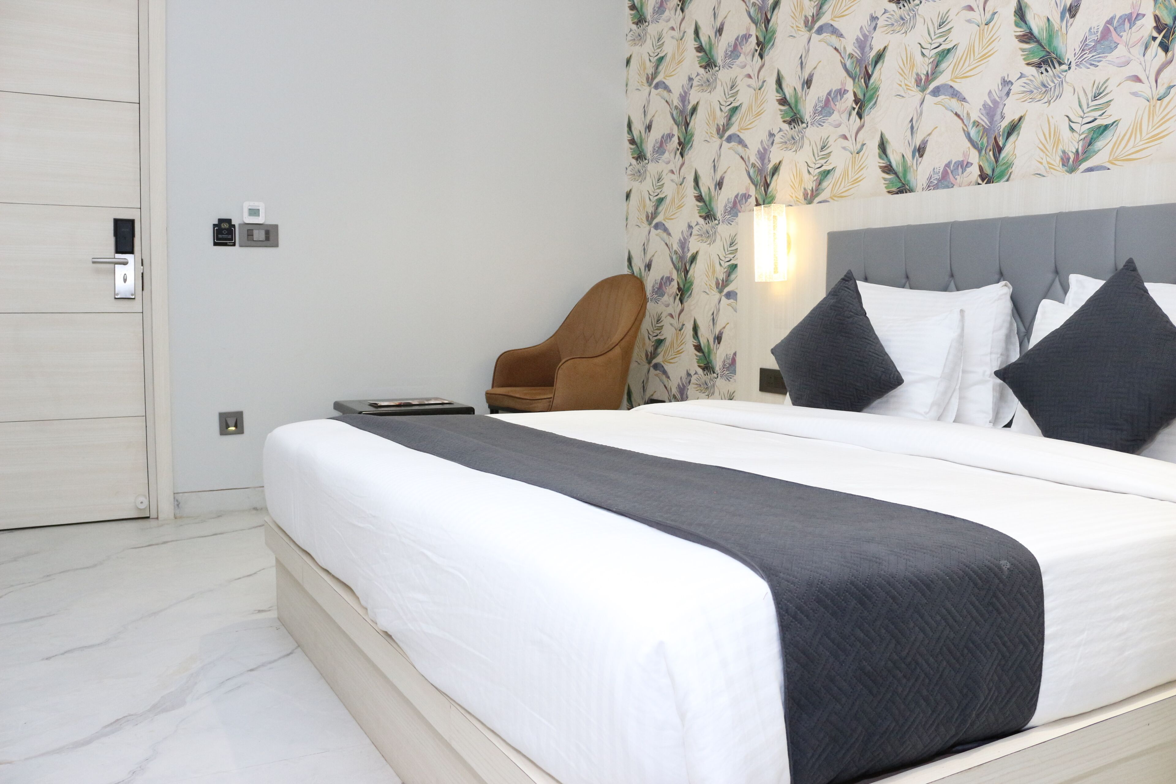 A neatly made bed with white and gray bedding, a brown chair, and a wall with a floral pattern.