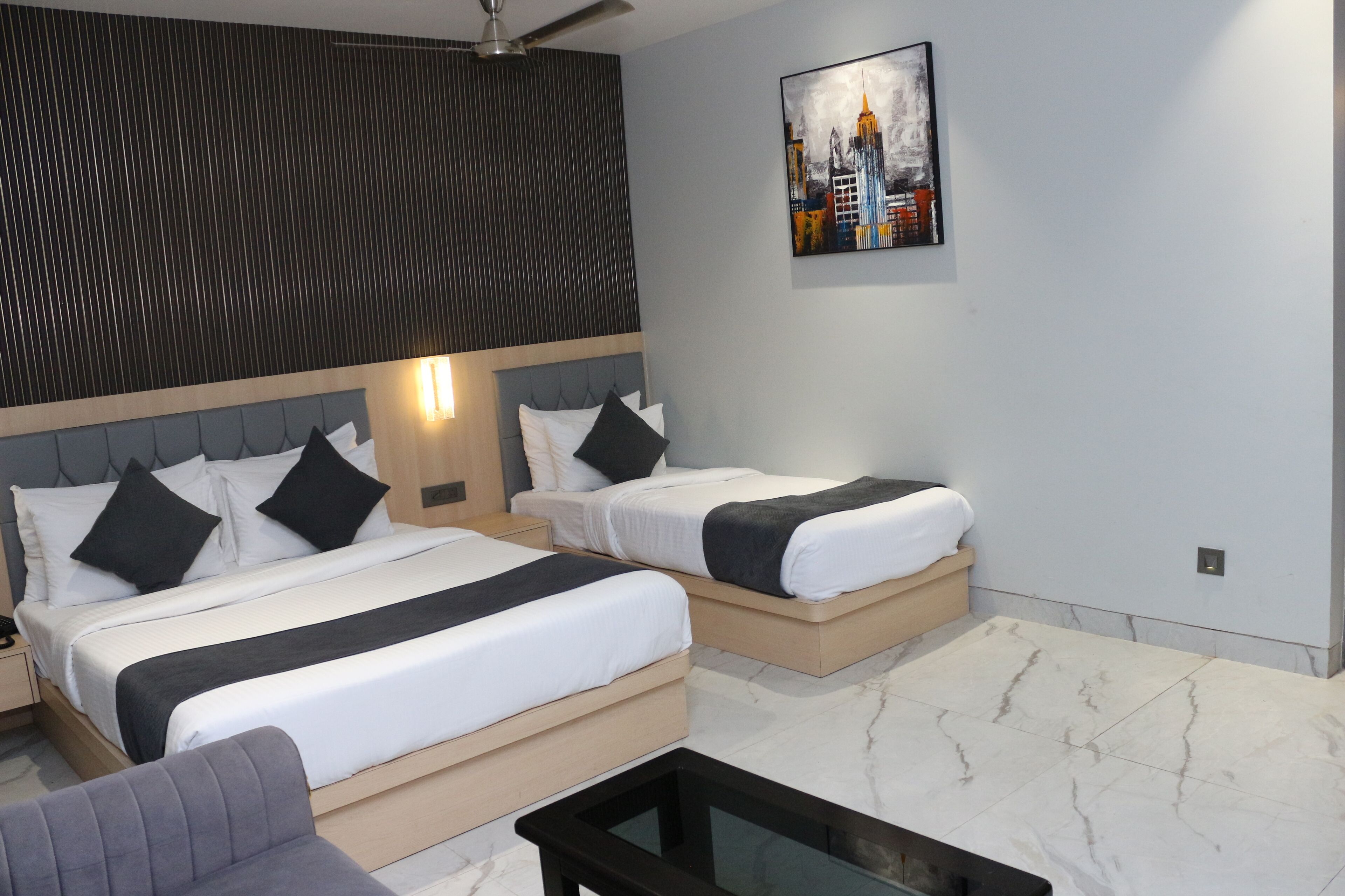 A hotel room with two beds, a sofa, a coffee table, a ceiling fan, and a cityscape painting on the wall.