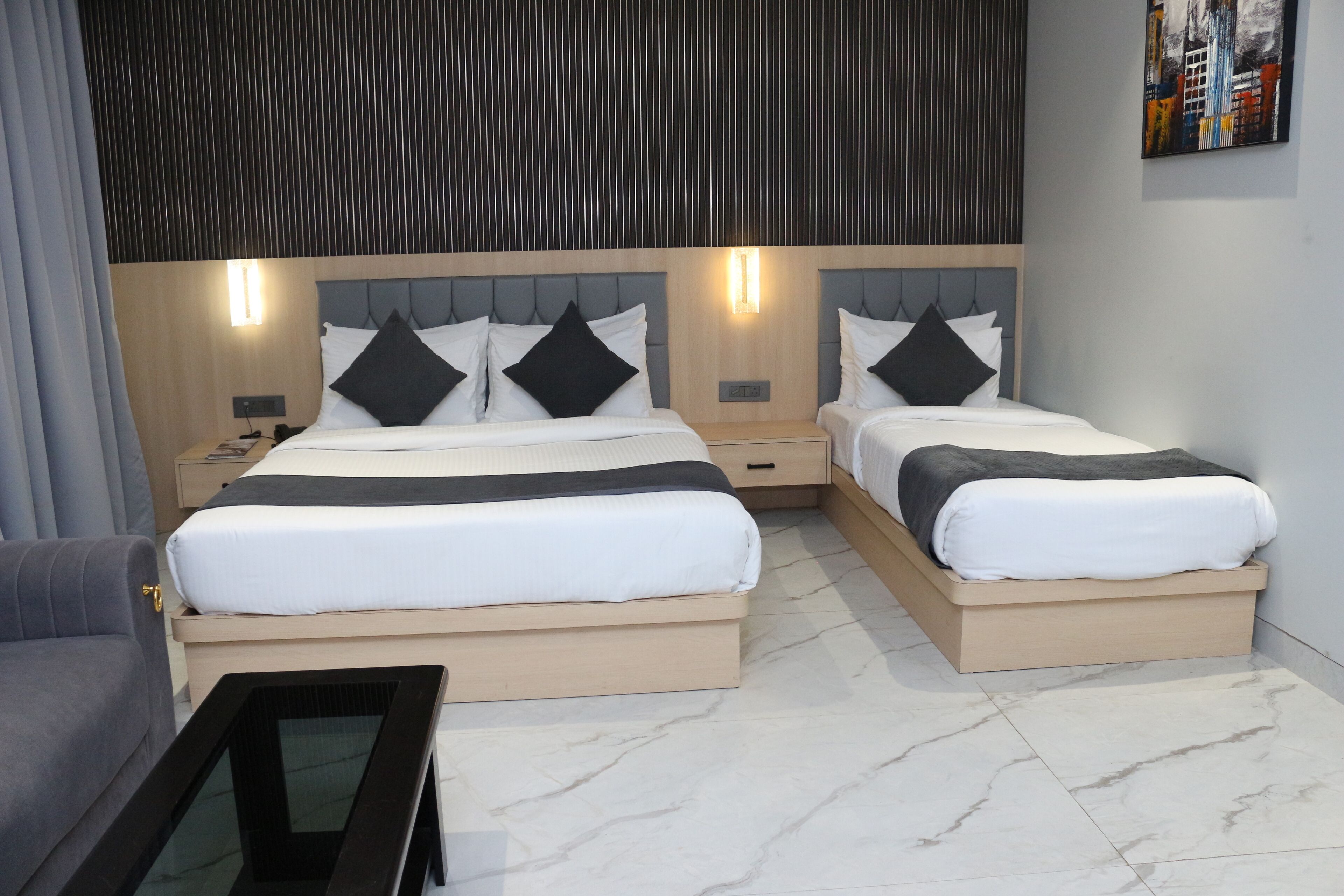 A modern hotel room with two beds, a sofa, a nightstand, and a wall-mounted artwork.