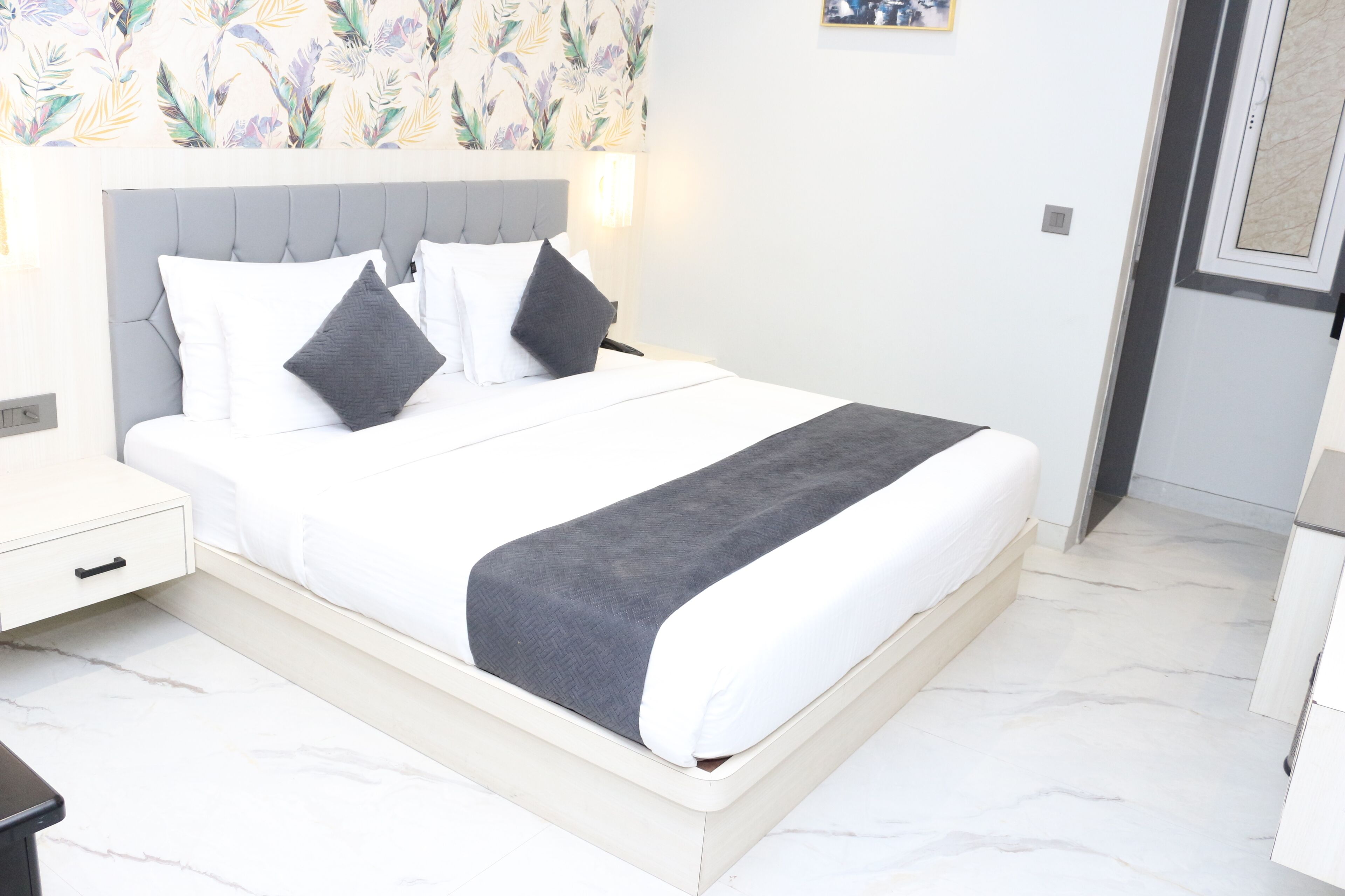 A neatly made bed with a grey headboard and white bedding, a bedside table with a drawer, and a wall with a floral wallpaper pattern.