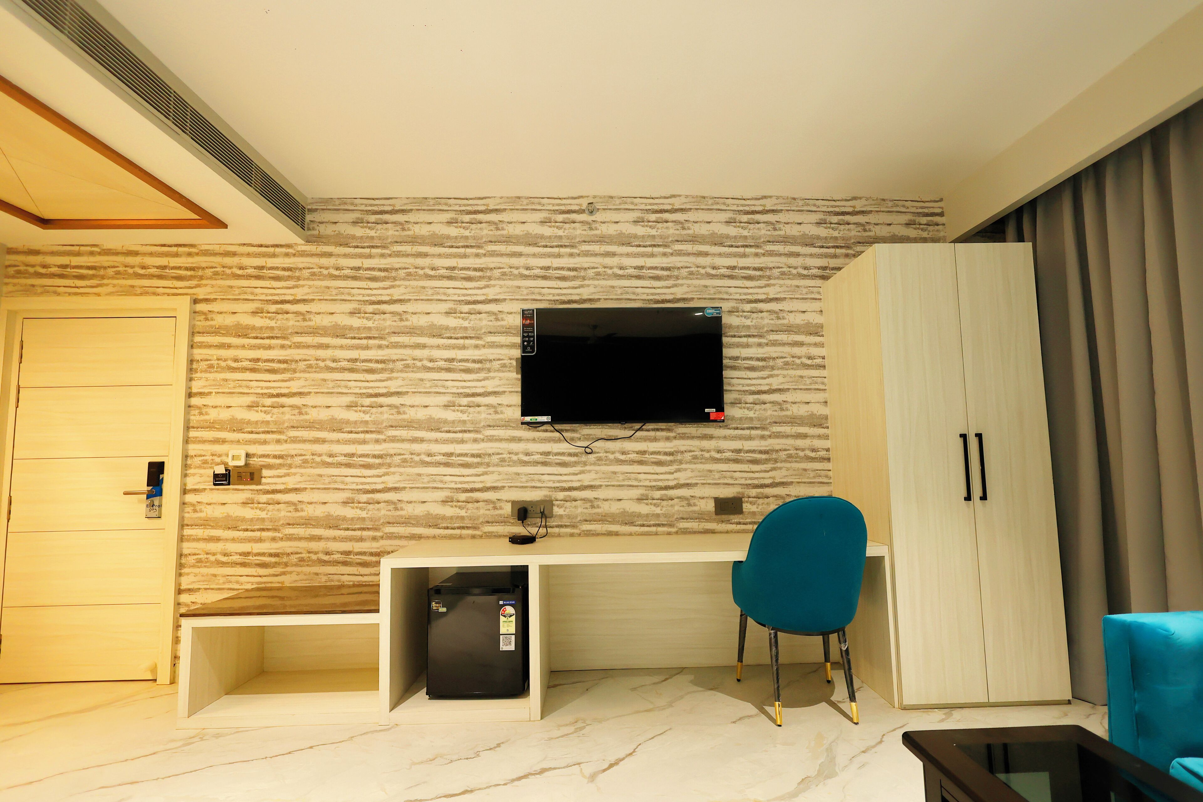 Une chambre moderne dotée d’un téléviseur à écran plat fixé à un mur texturé, d’un bureau avec une chaise, d’une armoire et d’un petit réfrigérateur.