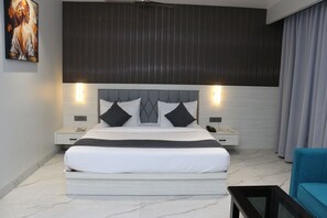 A modern bedroom with a large bed, a headboard with built-in lighting, a bedside table with a phone, and a blue chair. - Hotel Sankalp Banaras (Varanasi)
