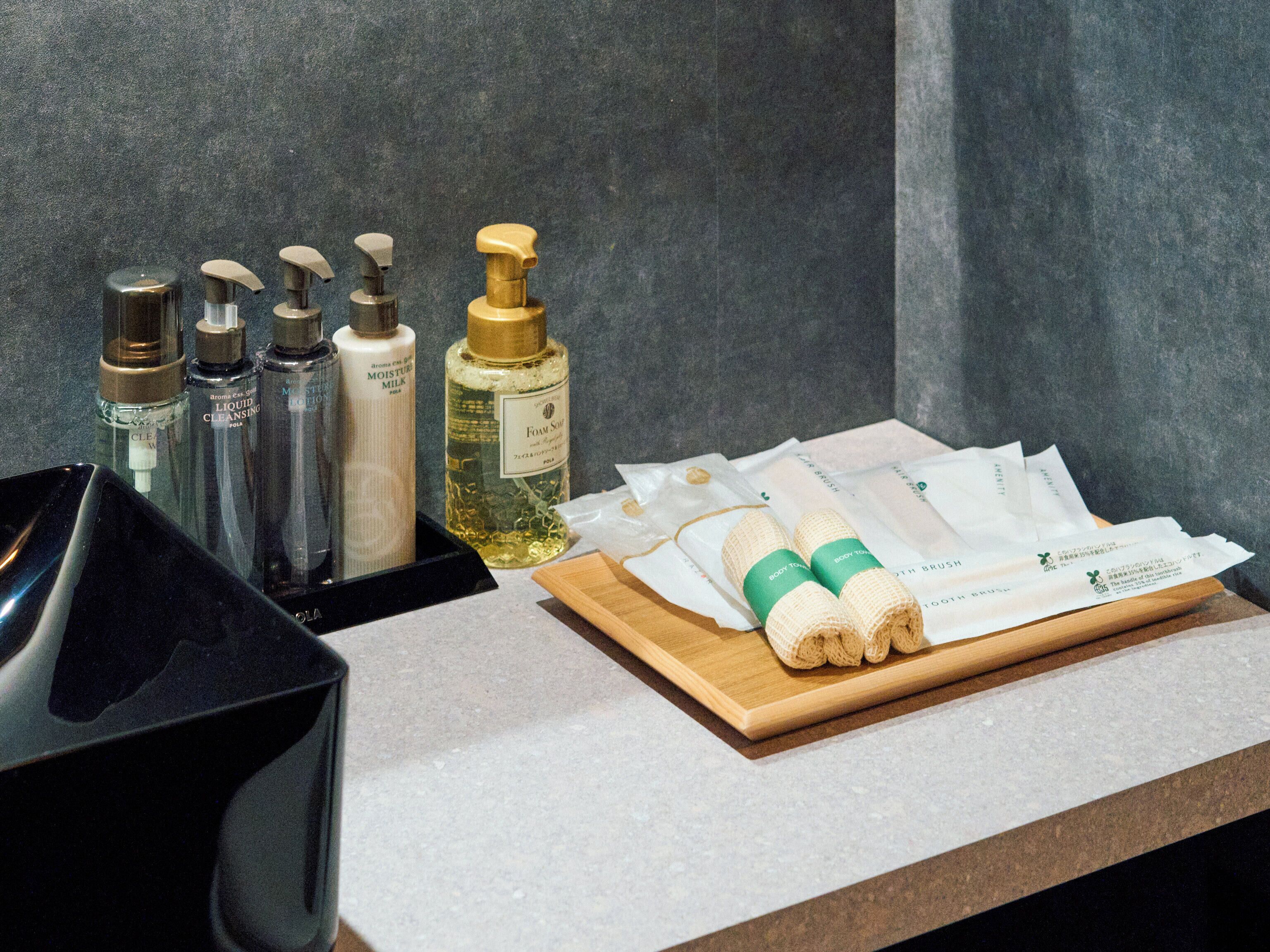 A bathroom counter with various toiletries including bottles of liquid soap, a bottle of foam soap, and a wooden tray with rolled towels and toiletries packaging.
