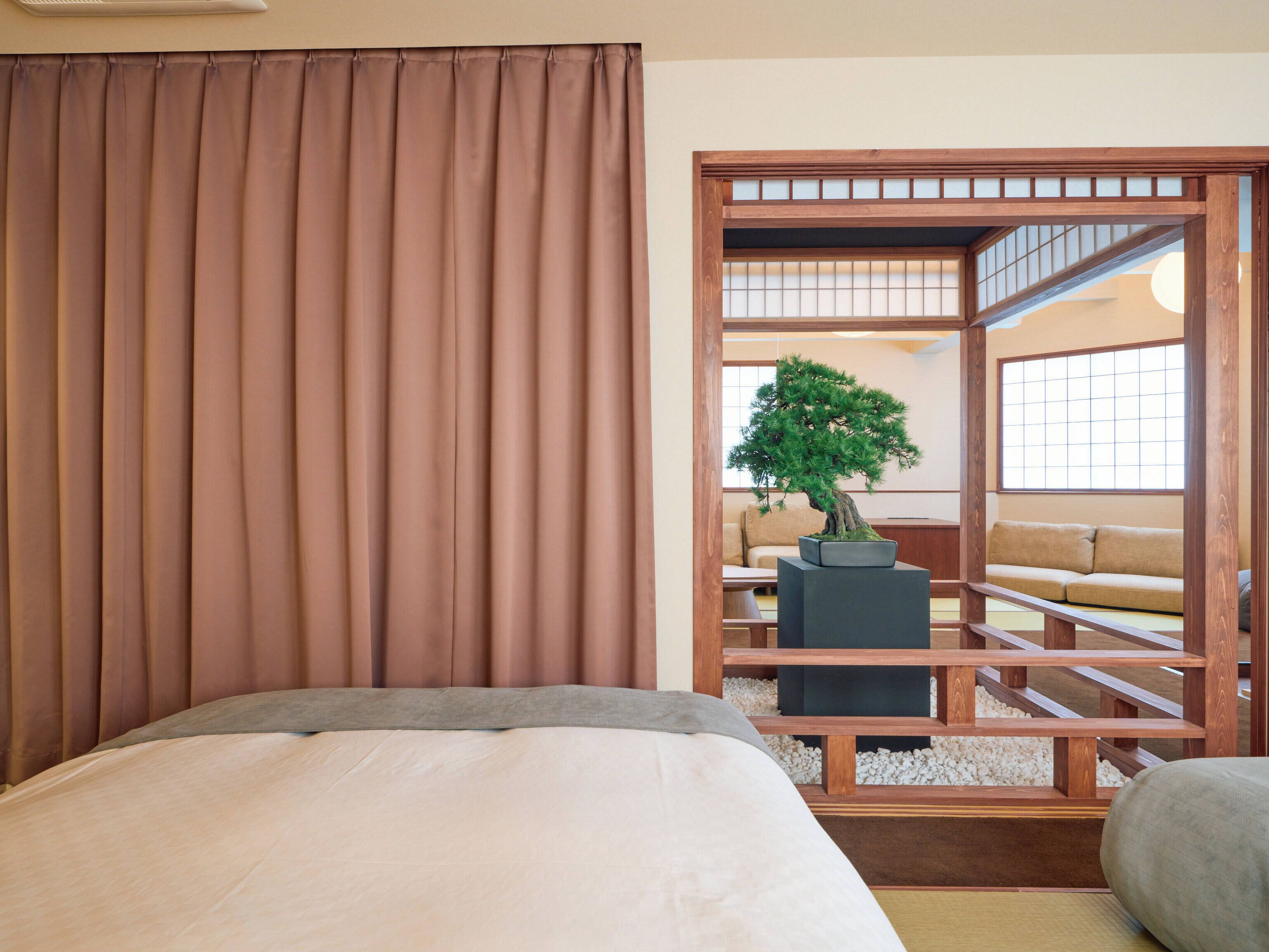 A room with a bed, a potted bonsai tree, a wooden partition, and a view of a traditional Japanese room.