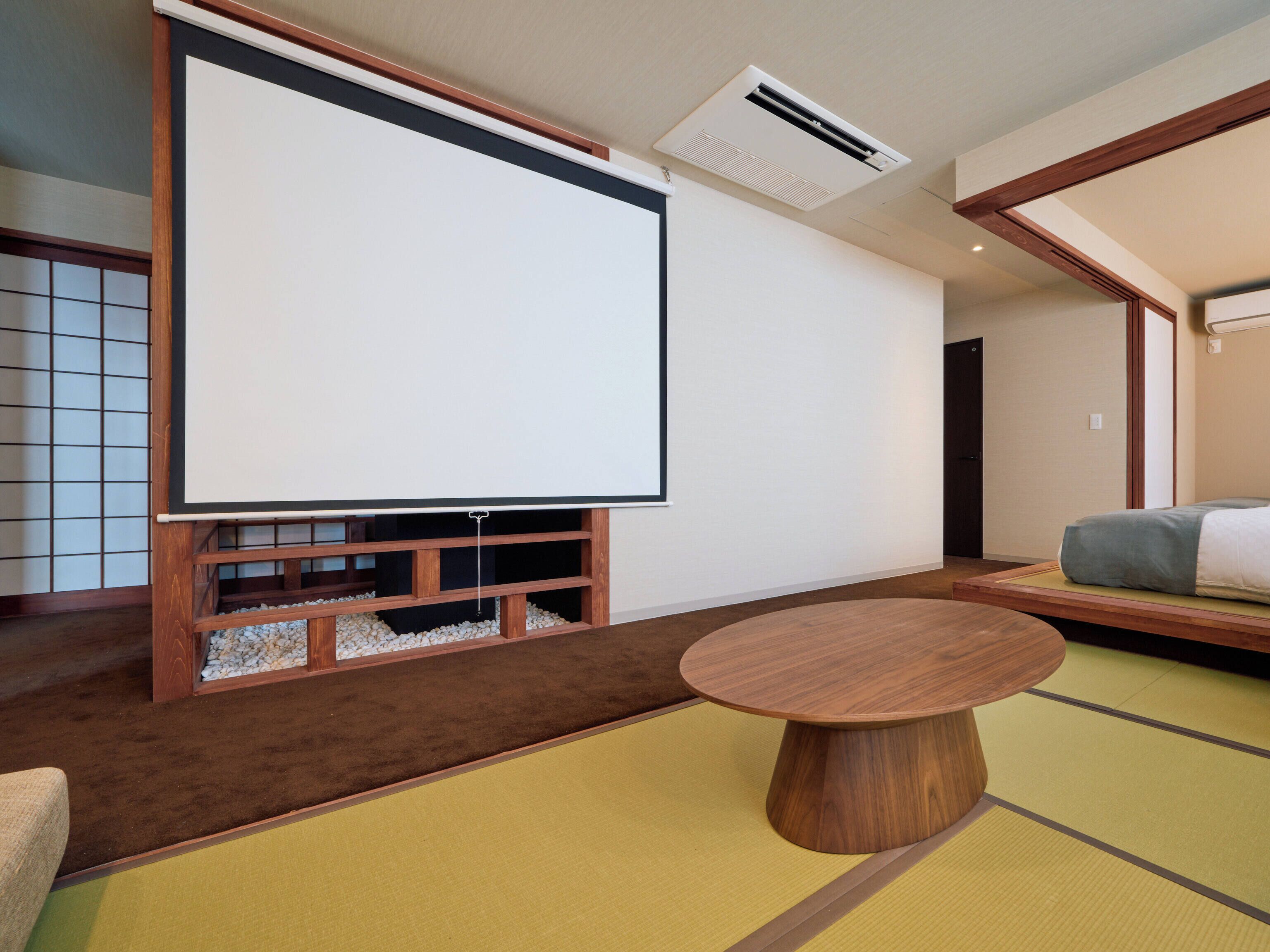 Una habitación con suelo de tatami, una mesa con tapete de tatami, un estante de madera bajo con una pantalla para proyector y una cama con futón.