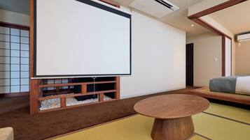 Una habitación con suelo de tatami, una mesa con tapete de tatami, un estante de madera bajo con una pantalla para proyector y una cama con futón.