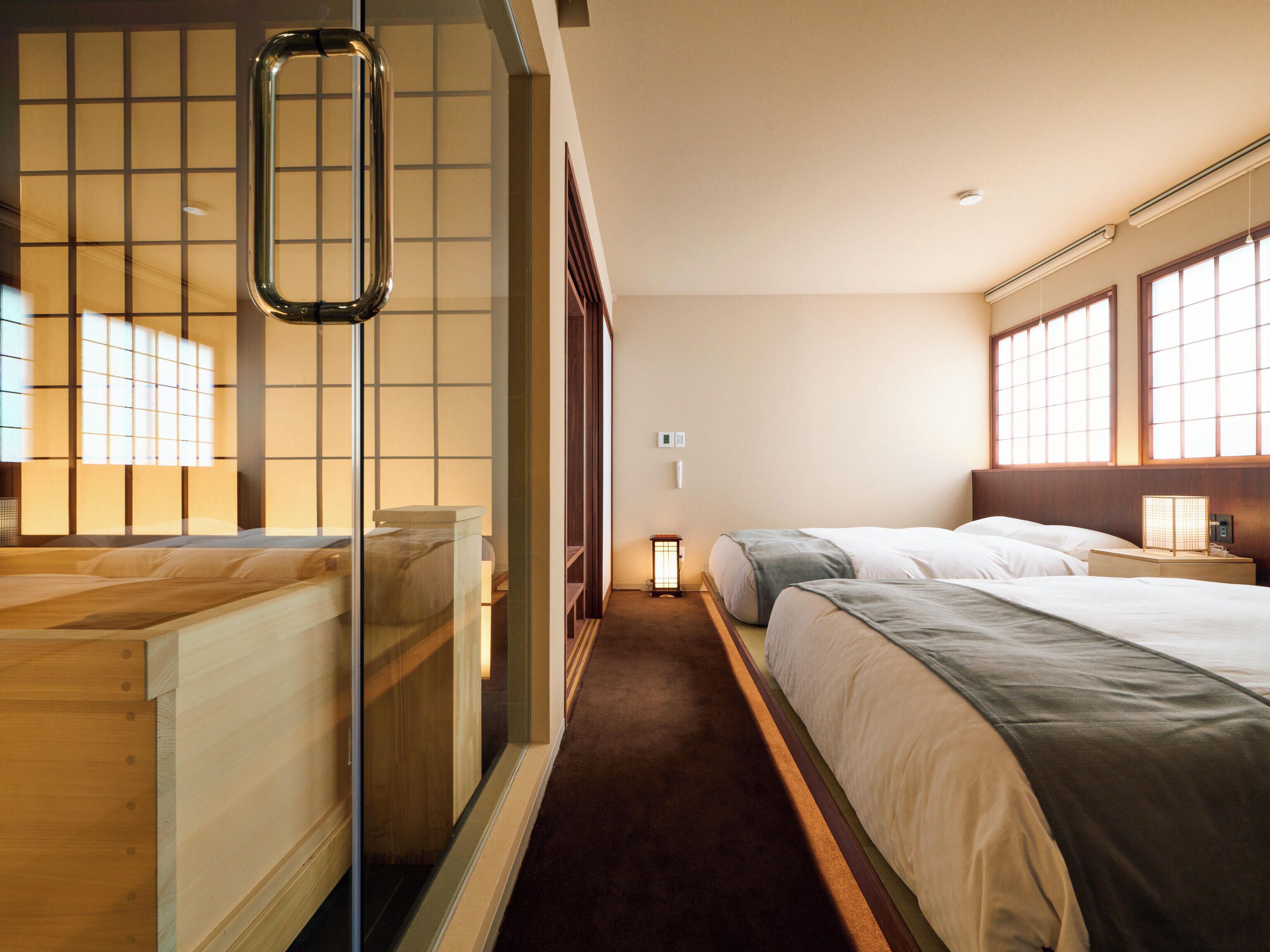 A hotel room with two beds, a sliding shoji screen, a small table with a lamp, and a view of the outside through large windows.