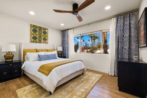 Une chambre avec un lit, un ventilateur de plafond, une fenêtre donnant sur des palmiers et une table de chevet avec une lampe.