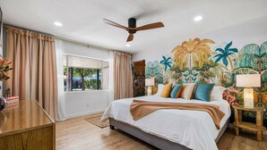Une chambre avec un grand lit, un ventilateur de plafond, une fenêtre avec des rideaux et une fresque murale sur le thème des tropiques.