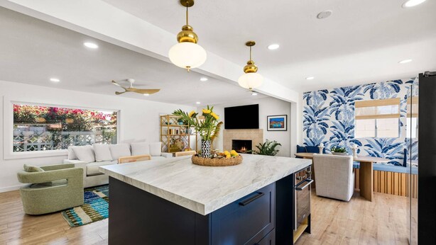 Une cuisine moderne dotée d’un îlot central, un coin repas avec un mur orné d’un motif bleu, et un salon avec une grande fenêtre donnant sur un jardin.