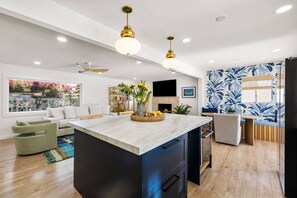 Une cuisine moderne dotée d’un îlot central, un coin repas avec un mur orné d’un motif bleu, et un salon avec une grande fenêtre donnant sur un jardin.