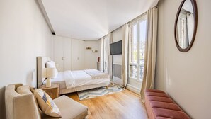 Un dormitorio con cama, mesita de noche con lámpara, una silla, un banco rojo, un ventanal con cortinas y un televisor instalado en la pared.