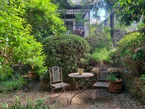 Une terrasse de jardin avec une table et des chaises en métal, entourée d’un feuillage dense et d’un mur de pierre.