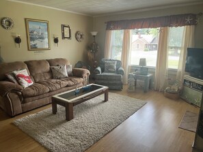 A living room with a brown sofa, a coffee table, a patterned armchair, a television, and a window with curtains.