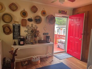 Une pièce chaleureuse avec une porte rouge, une console blanche ornée de divers objets, un mur décoré de tournesols et une pancarte indiquant « Bienvenue, amis », et une vue sur une maison à l’extérieur.