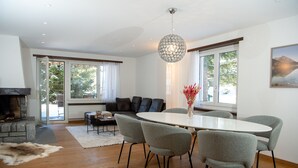 Ein modernes Wohnzimmer mit Kamin, einem Esstisch mit Stühlen, einem großen Fenster und einer Hängeleuchte.