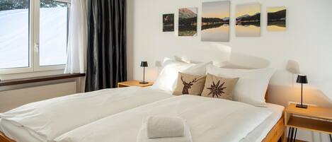 A hotel room with a large bed, two bedside tables with lamps, a window with curtains, and three landscape paintings on the wall.