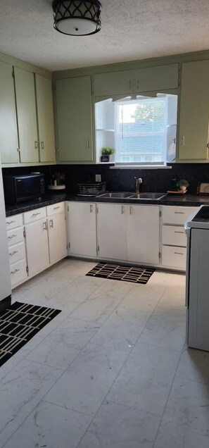 A kitchen with light green cabinets, a black countertop, a window with blinds, and a microwave.