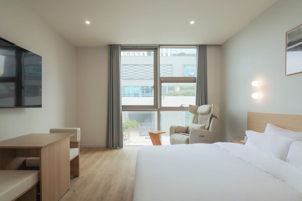 A modern hotel room with a large bed, a desk, a chair, and a view of the outside through large windows. - Anook Hotel Sejong (Sejong)