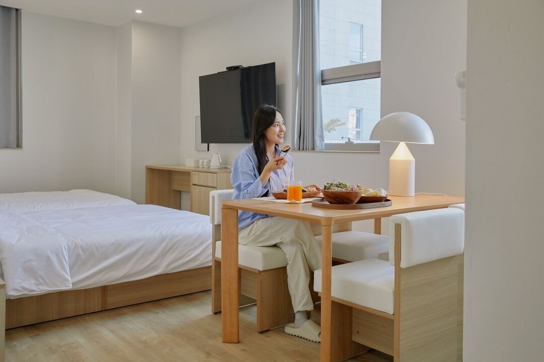 A modern hotel room with a bed, a dining table with breakfast, a TV, and a window with a view.