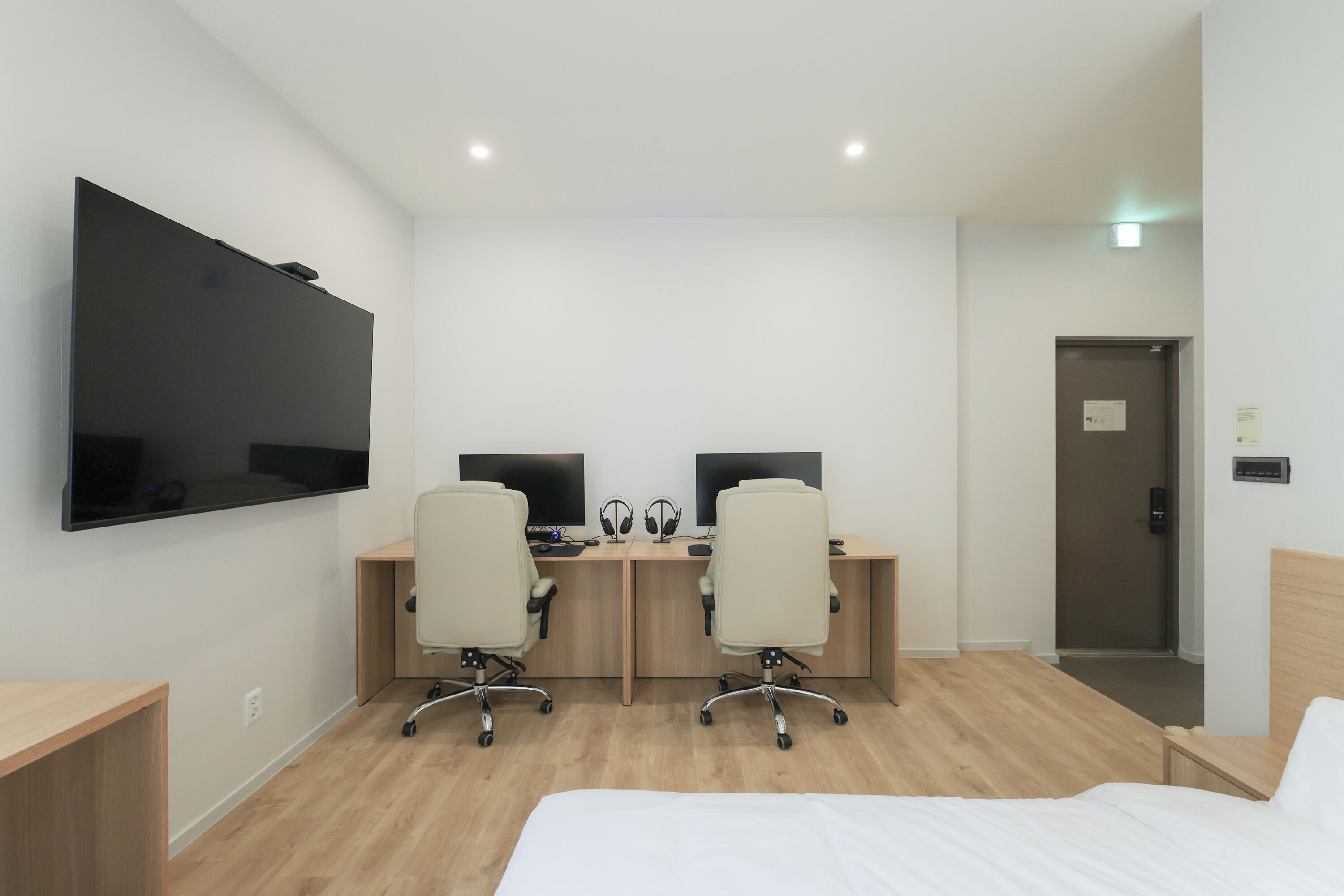 Habitación de hotel con un televisor de pantalla plana grande montado en la pared, dos escritorios con computadoras y audífonos, una cama con ropa blanca y piso de madera.