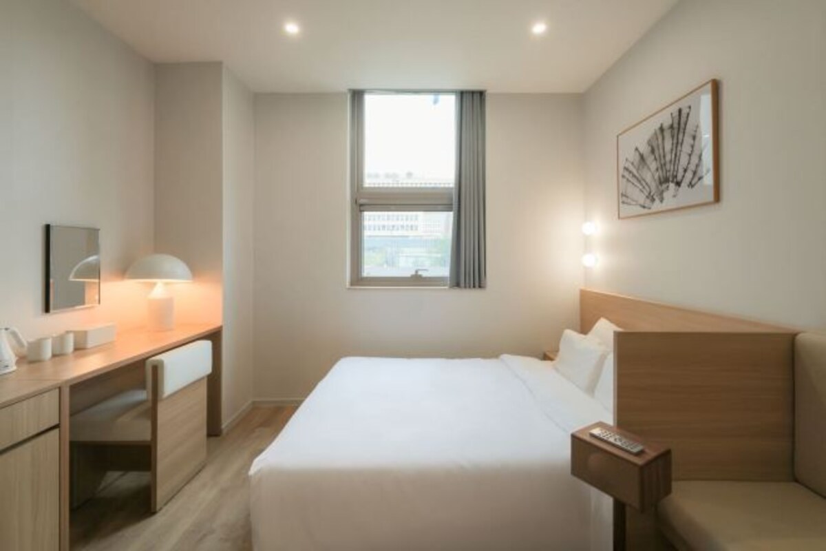 A modern hotel room with a bed, bedside table, desk with chair, and a window with curtains.