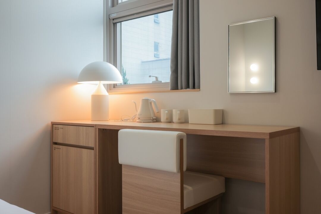 Una habitación moderna con un escritorio de madera, una silla blanca, una lámpara, un hervidor y un espejo.