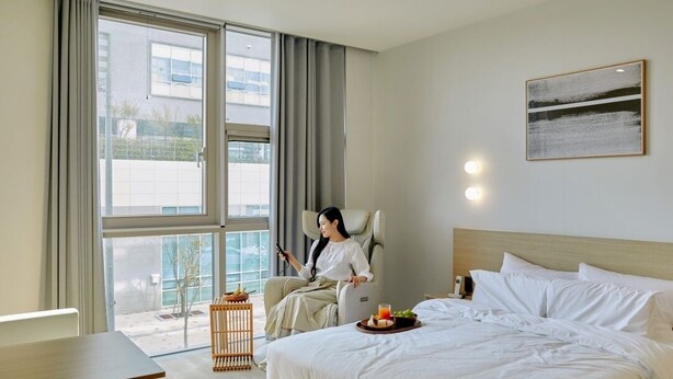 A modern hotel room with a large bed, a sitting area with a chair, a small table, and a view of the outside through a large window. - Anook Hotel Sejong (Sejong)