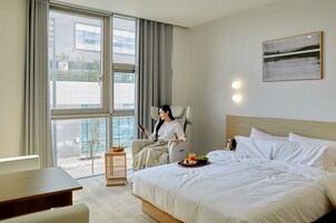 Una habitación de hotel moderna con una cama grande, una zona de estar con una silla, una mesita y vistas al exterior a través de un ventanal amplio.