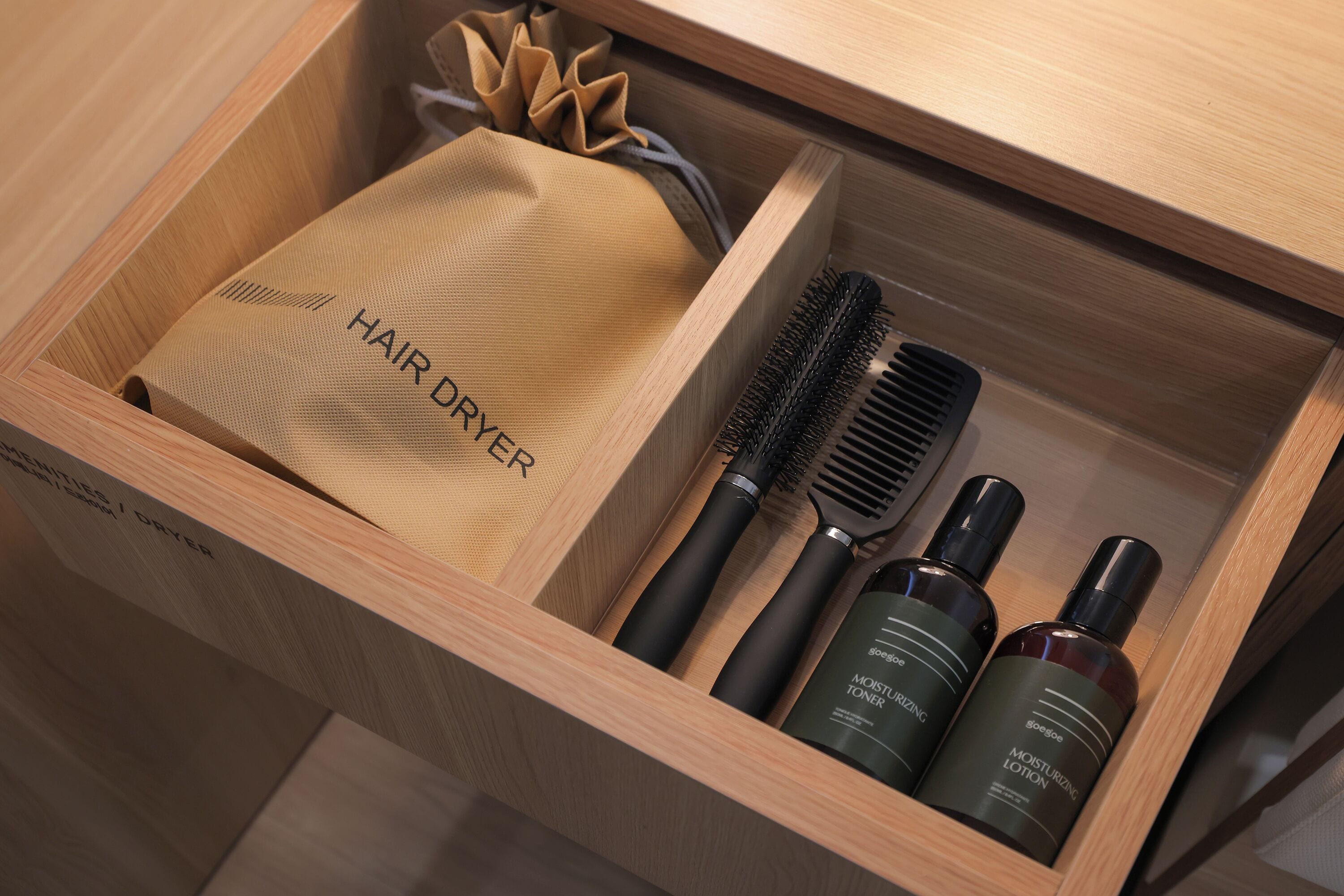 A wooden drawer containing a hair dryer, two hairbrushes, and two bottles of hair care products.