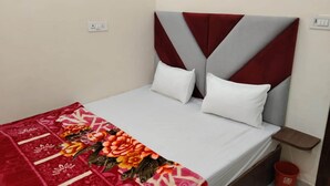 Een eenpersoonsbed met een rood-wit gebloemd beddegoed, een grijs gestoffeerd hoofdbord en witte kussens. Er staat een klein houten nachtkastje met een rode emmer naast het bed.