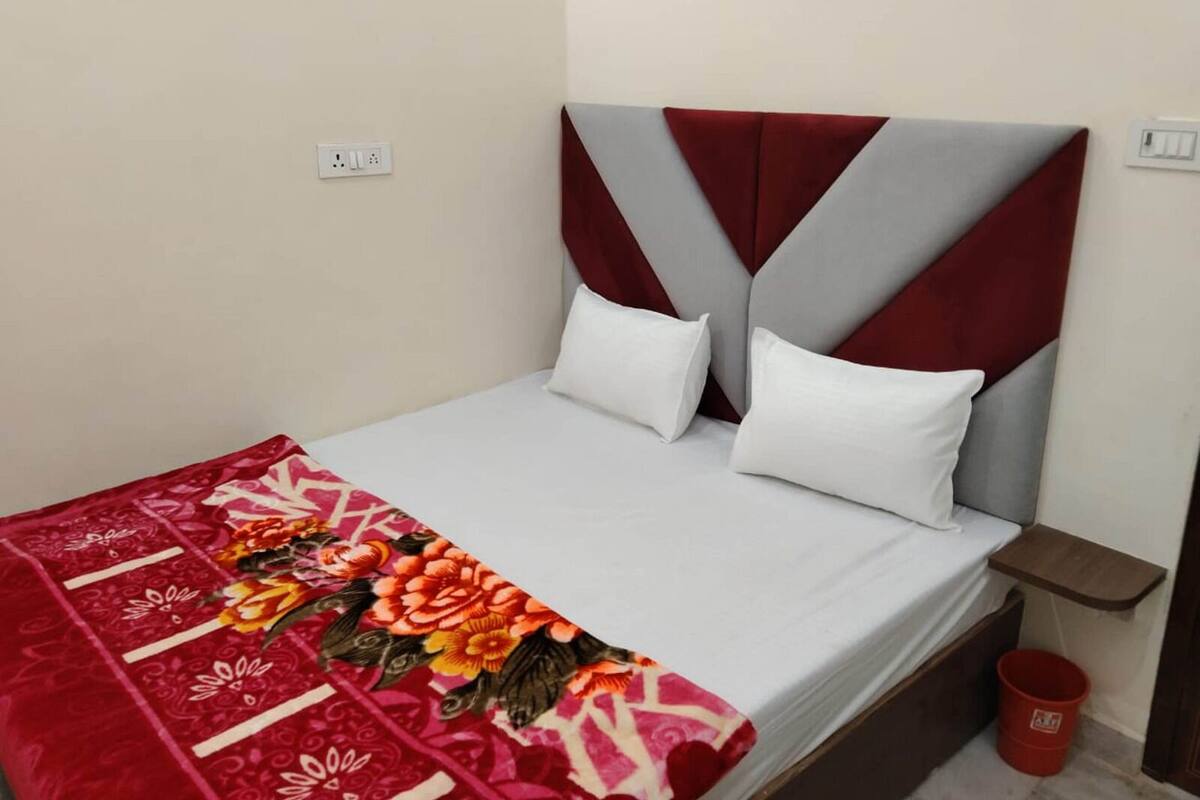 A single bed with a red and white floral bedspread, a grey upholstered headboard, and white pillows. There is a small wooden nightstand with a red bucket beside the bed.