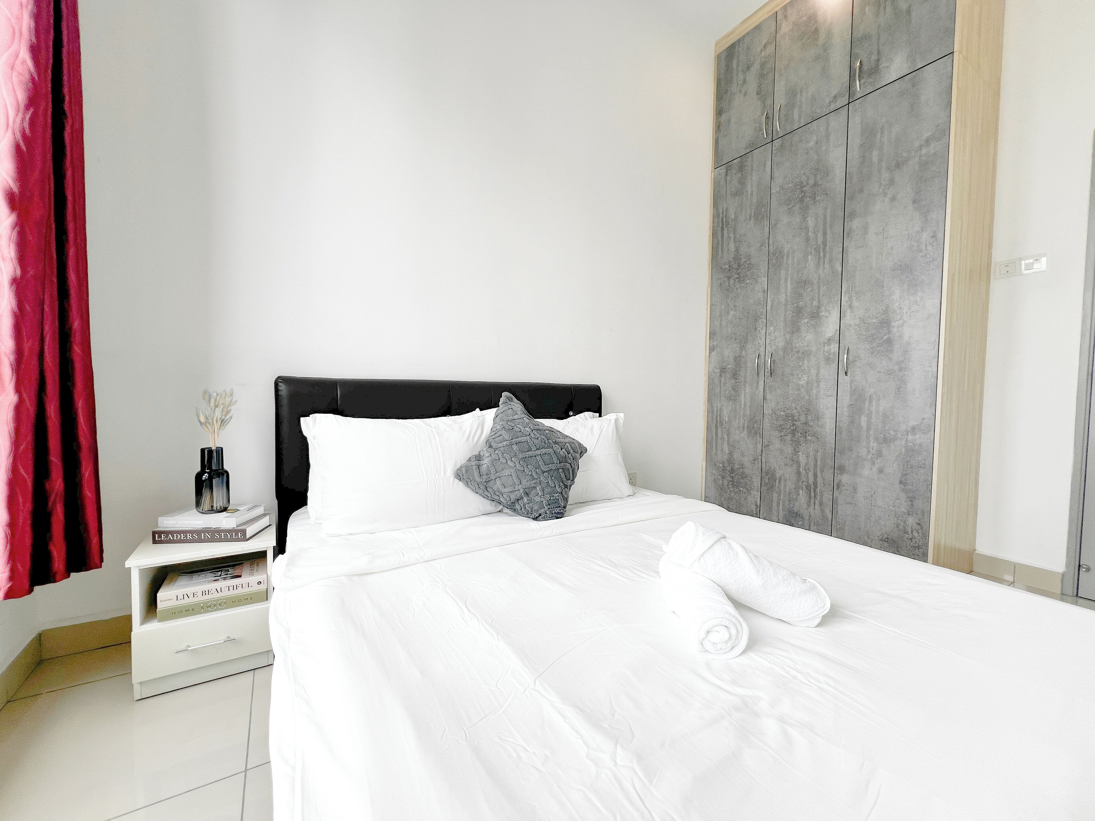 Un lit bien fait, avec du linge de lit blanc, un oreiller gris et une serviette enroulée. On aperçoit une table de chevet blanche sur laquelle repose un livre intitulé « VIVEZ PLEINEMENT ». Une armoire grise est adossée au mur.