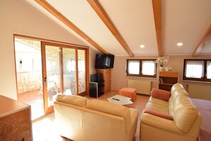 Relax in this spacious living area with a high ceiling and exposed beams, featuring a sofa, flat-screen TV, and sliding glass doors leading to an outdoor patio.